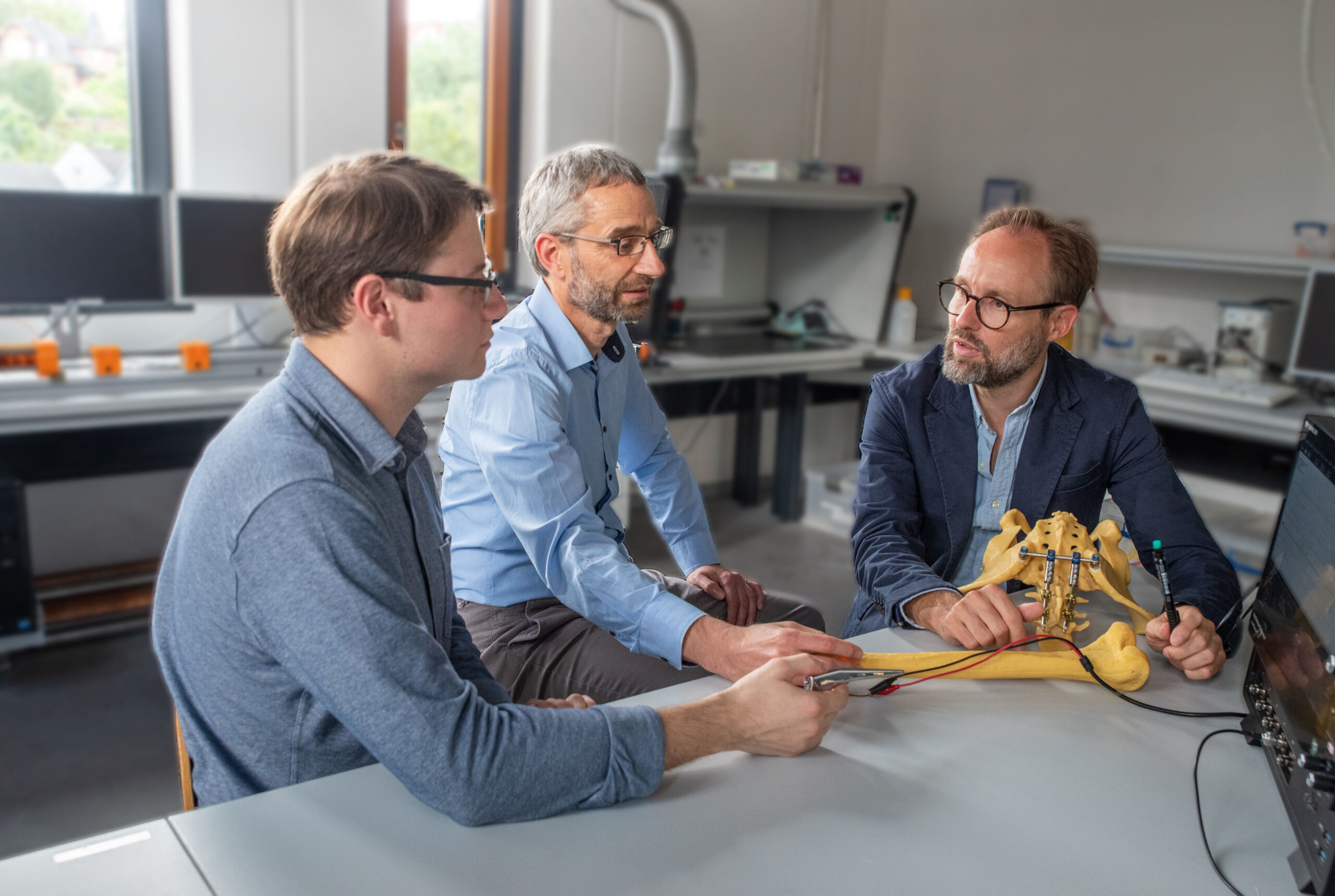 In einem Labor, in dem emsig geforscht wird, sitzen drei Männer um einen Tisch herum und untersuchen ein Roboterhundemodell. Einer hält ein Steuergerät in der Hand, ein anderer gestikuliert lebhaft auf das Modell, während der dritte aufmerksam zusieht. Computer und Geräte füllen den Hintergrund, als ob sie von einem aufmerksamen Coburger Studenten beaufsichtigt würden. at Hochschule Coburg