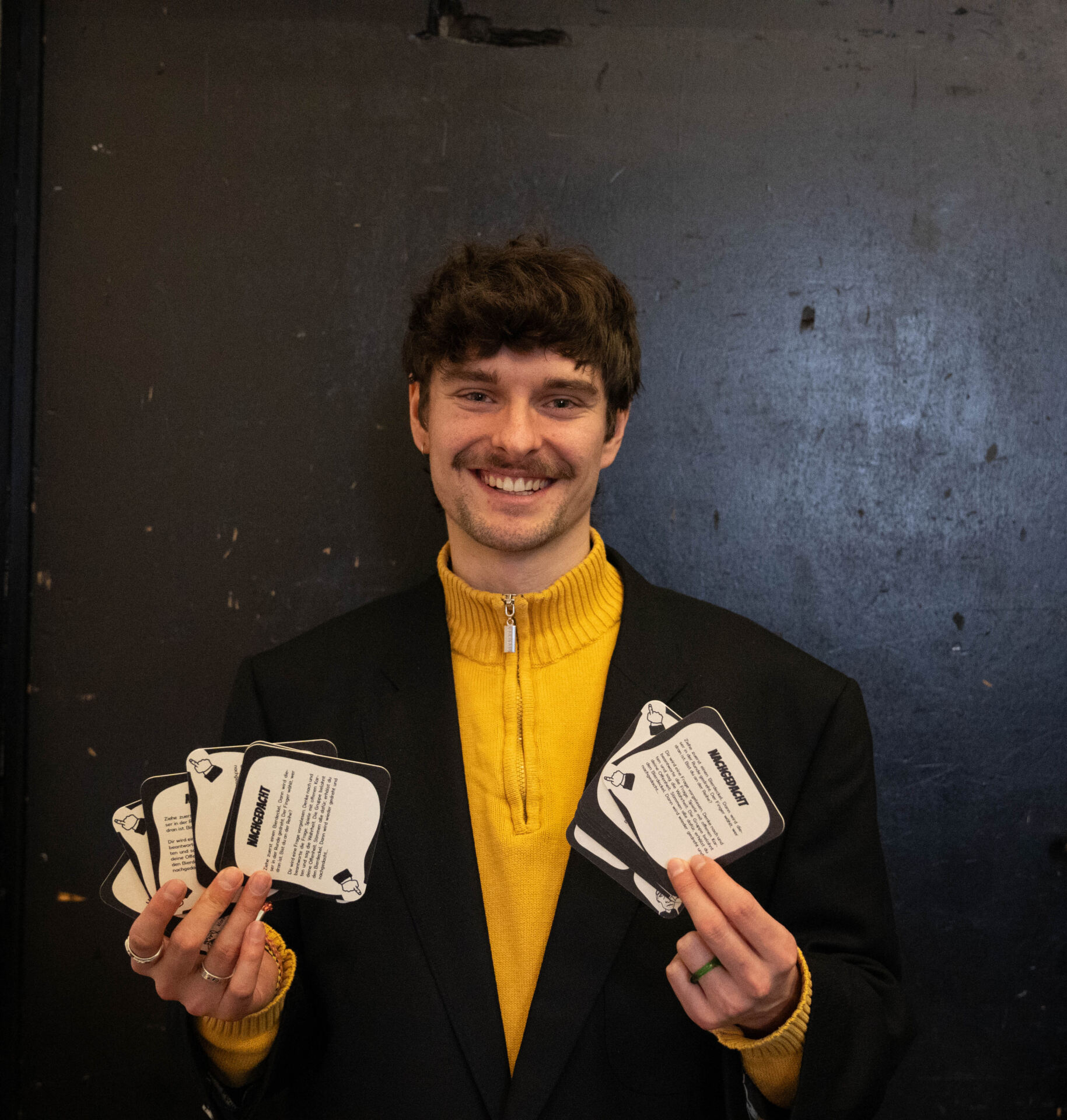 Eine Person mit lockigem Haar und Schnurrbart lächelt und hält mehrere Karten in der Hand, was den Design-Aktivismus verkörpert. Sie trägt eine schwarze Jacke über einem leuchtend gelben Pullover. Der Hintergrund ist eine dunkle, strukturierte Wand, perfekt, um eine Diskussion anzuregen. at Hochschule Coburg