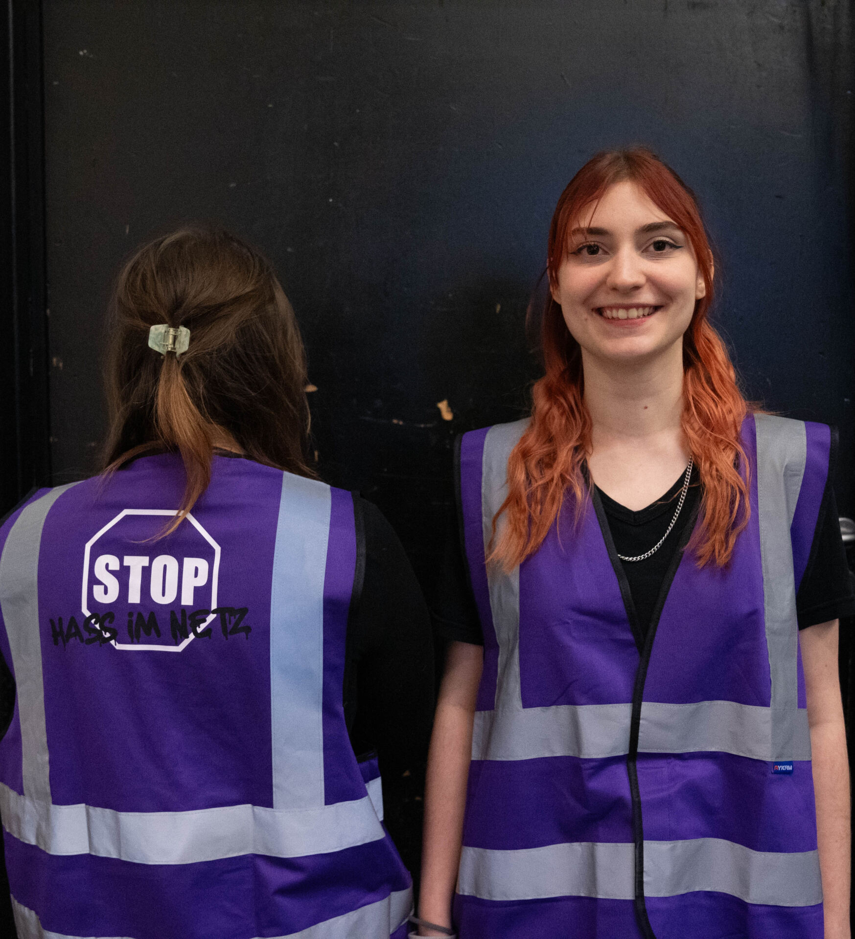 Zwei Personen in lila Westen mit Reflektorstreifen stehen vor einem dunklen Hintergrund. Die rechte Person lächelt in die Kamera, während auf der Rückseite der Weste der anderen Person „STOP“ und „Hass im Netz“ zu sehen sind, was ihre Teilnahme an einem Studierenden-Projekt mit dem Schwerpunkt Design-Aktivismus unterstreicht. at Hochschule Coburg