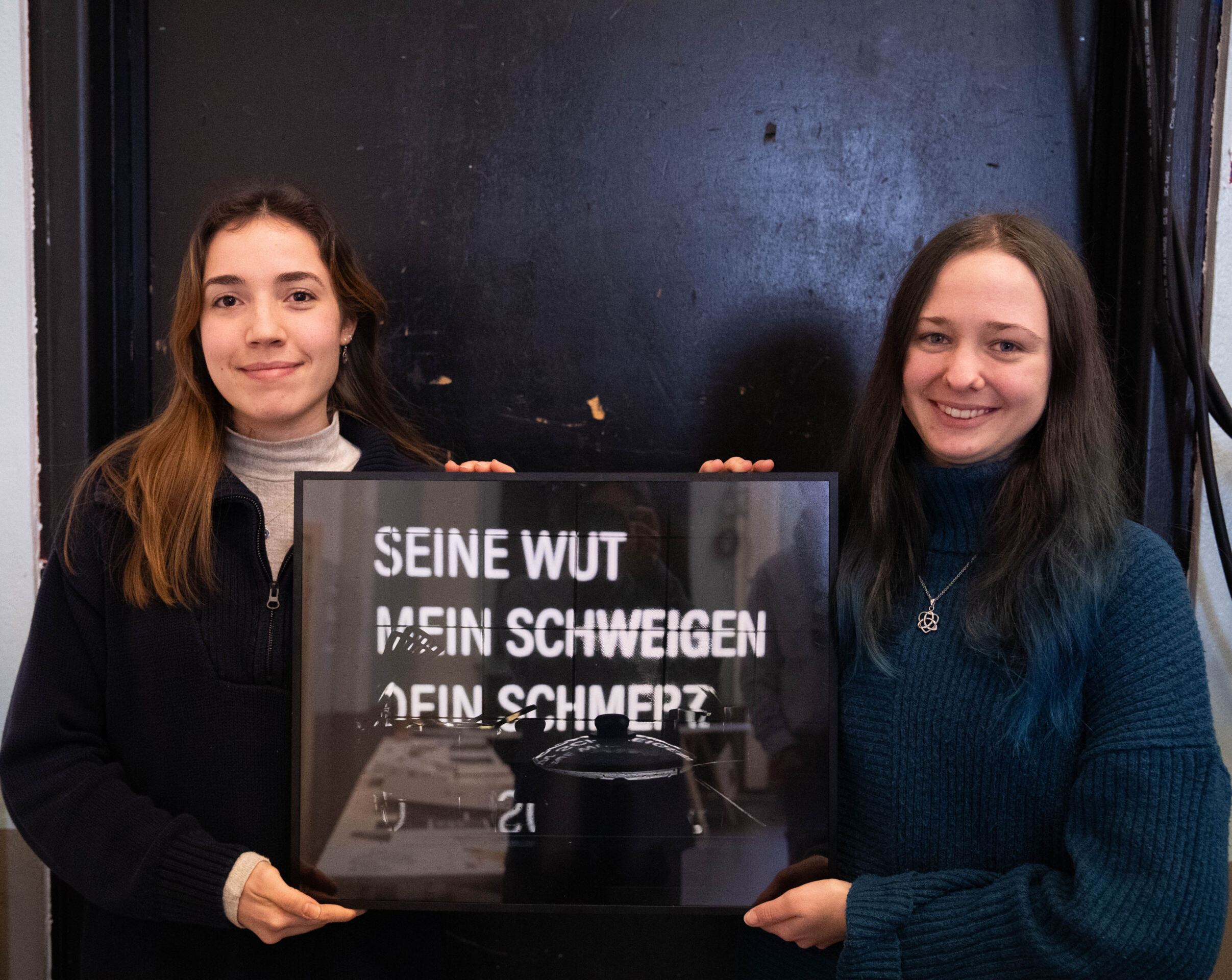 Zwei Frauen stehen Seite an Seite und halten ein gerahmtes Bild mit dem deutschen Text „SEINE WUT, MEIN SCHWEIGEN, DEIN SCHMERZ“ in kräftigen weißen Buchstaben auf dunklem Hintergrund. Mit einem warmen Lächeln in lässigen Pullovern präsentieren sie dieses Stück Design-Aktivismus aus einem inspirierenden Studierenden-Projekt. at Hochschule Coburg