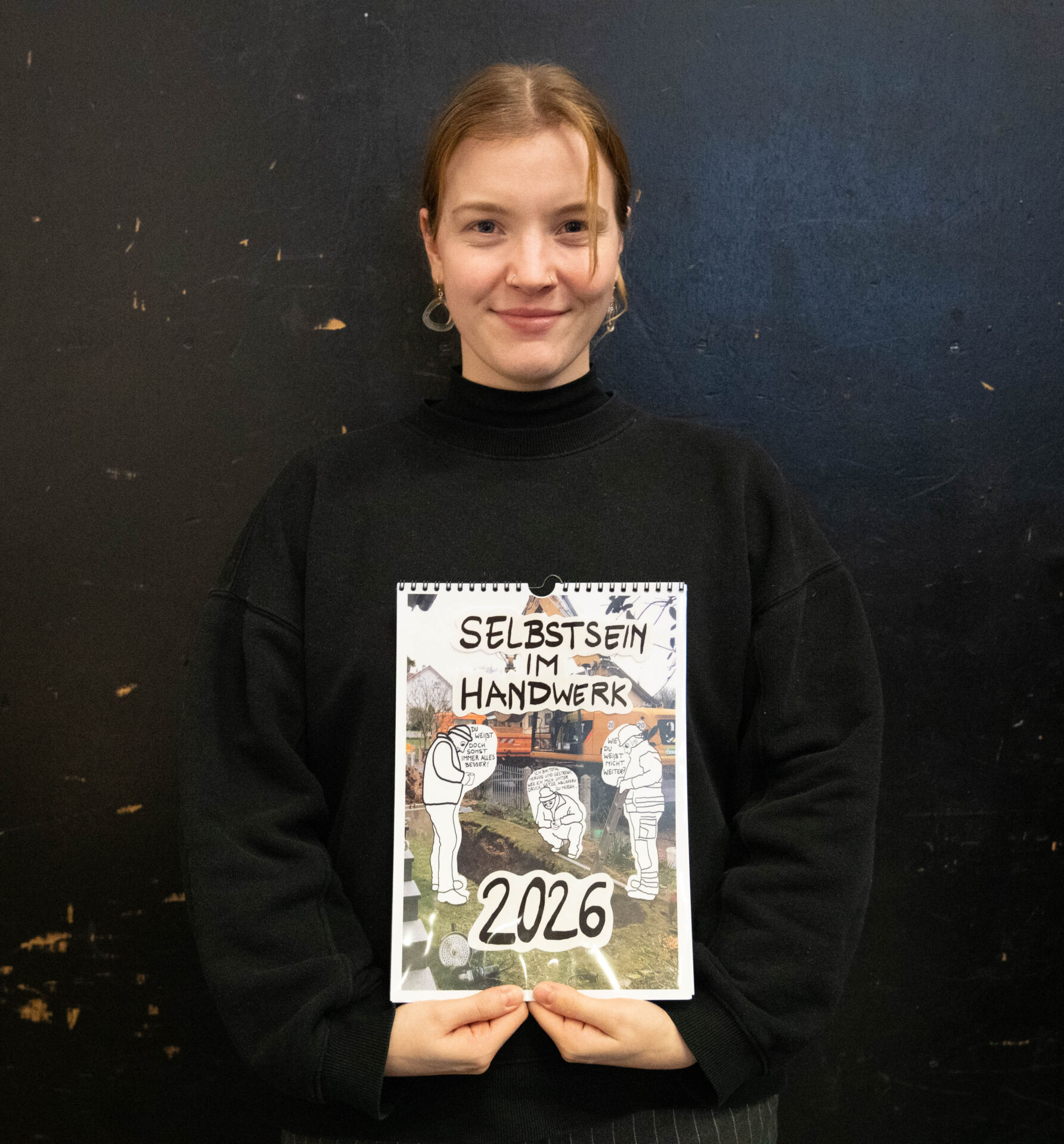 Eine Person in einem schwarzen Pullover hält einen Kalender mit illustrierten Figuren und dem Text „Selbstsein im Handwerk 2026“. Der Kalender, ein Ergebnis eines Studierenden-Projekts mit dem Schwerpunkt Design-Aktivismus, steht vor einem dunklen, schlichten Hintergrund. at Hochschule Coburg