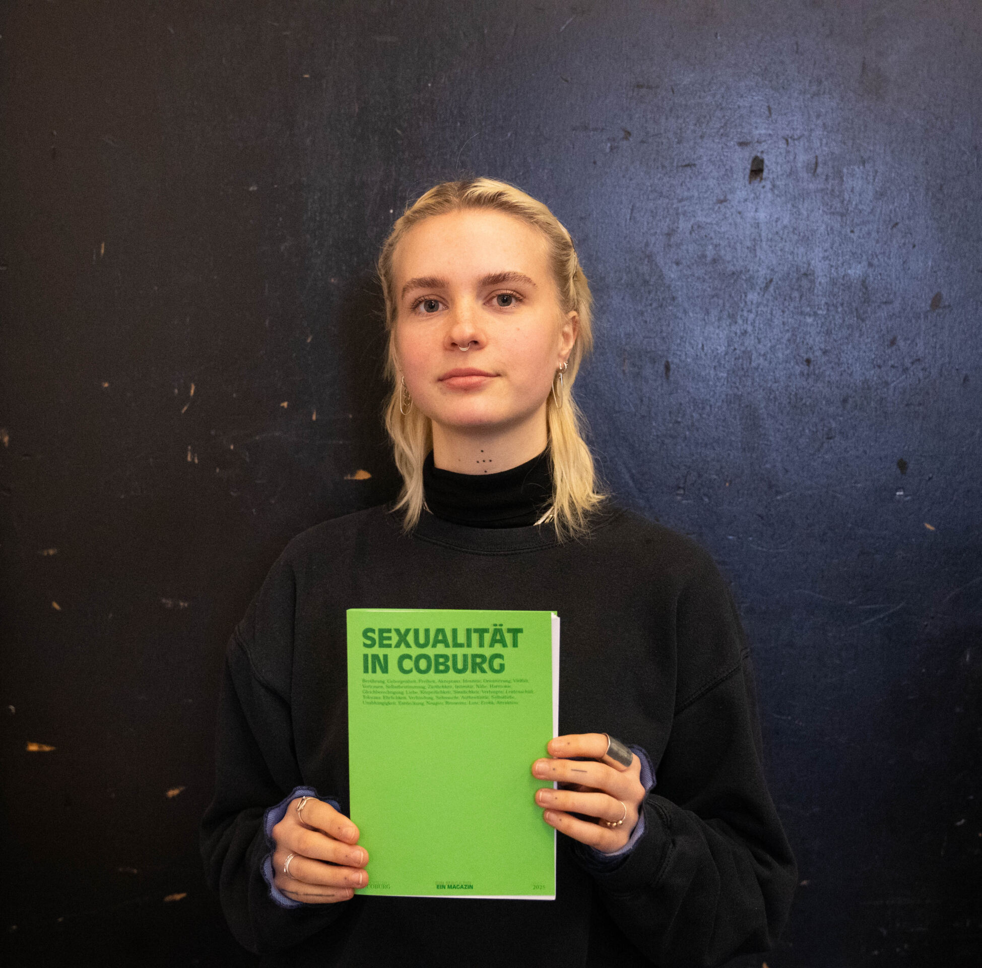 Eine Person mit langen blonden Haaren und schwarzem Pullover hält vor einem dunklen, strukturierten Hintergrund ein grünes Buch mit dem Titel „SEXUALITÄT IN COBURG“. Im Rahmen eines Studierendenprojekts zum Thema Design-Aktivismus blickt sie direkt in die Kamera und lädt zu einer Diskussion über das Thema ein. at Hochschule Coburg