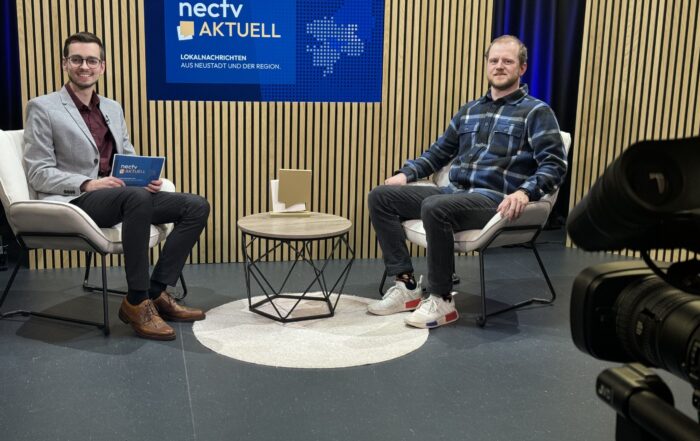 Zwei Menschen sitzen in einem Studio mit gemusterter Holzkulisse. Der eine hält ein Tablet in der Hand und trägt einen grauen Blazer, der andere ein kariertes Hemd und Turnschuhe. Zwischen ihnen steht ein kleiner Tisch unter dem Schild „nectv aktuell“ an der Wand. Eine Kamera fängt ihre Diskussion über ein innovatives Superkondensator-Projekt für das hr-Fernsehen ein. at Hochschule Coburg