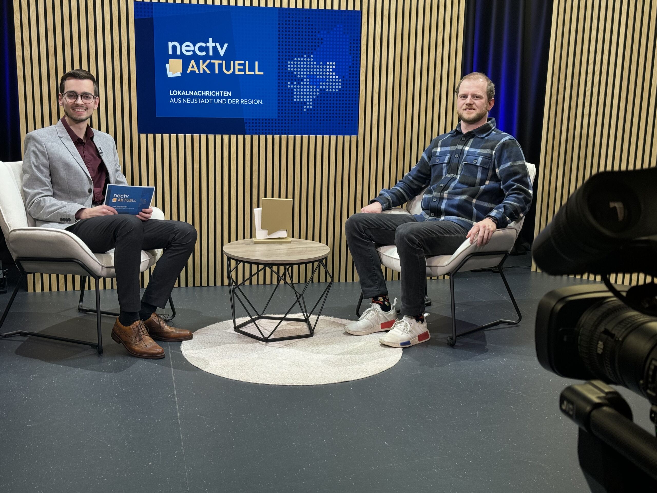 Zwei Menschen sitzen in einem Studio mit gemusterter Holzkulisse. Der eine hält ein Tablet in der Hand und trägt einen grauen Blazer, der andere ein kariertes Hemd und Turnschuhe. Zwischen ihnen steht ein kleiner Tisch unter dem Schild „nectv aktuell“ an der Wand. Eine Kamera fängt ihre Diskussion über ein innovatives Superkondensator-Projekt für das hr-Fernsehen ein. at Hochschule Coburg