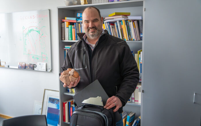 Eine lächelnde Person hält in einer Hand ein Modell eines menschlichen Gehirns und stützt die andere auf einen Rucksack. Sie verkörpert damit die Essenz von Produktivität. Sie steht vor einem Bücherregal voller Bücher und neben einem Whiteboard mit Diagrammen, vielleicht um Ideen für effizientere Homeoffice-Einrichtungen aufzuzeichnen. at Hochschule Coburg