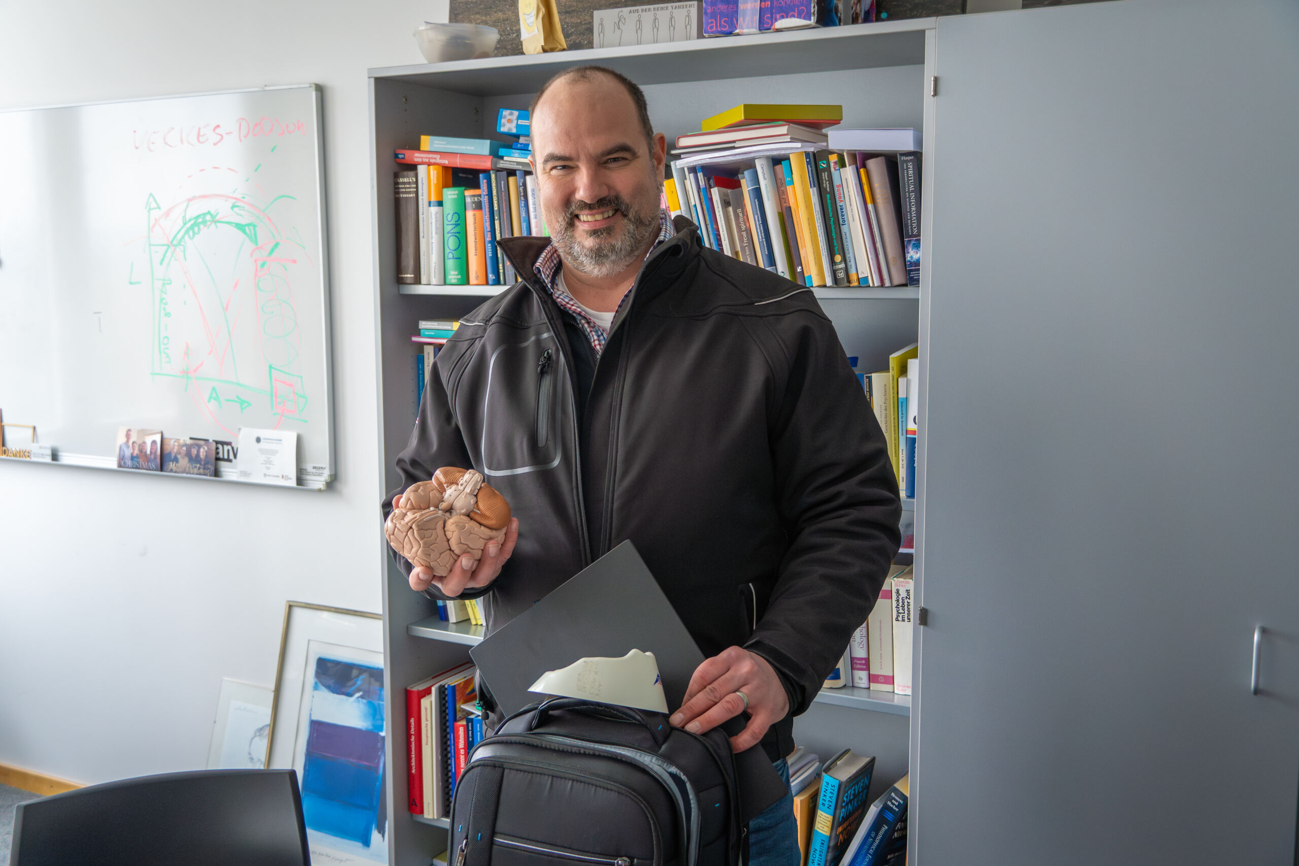 Eine lächelnde Person hält in einer Hand ein Modell eines menschlichen Gehirns und stützt die andere auf einen Rucksack. Sie verkörpert damit die Essenz von Produktivität. Sie steht vor einem Bücherregal voller Bücher und neben einem Whiteboard mit Diagrammen, vielleicht um Ideen für effizientere Homeoffice-Einrichtungen aufzuzeichnen. at Hochschule Coburg