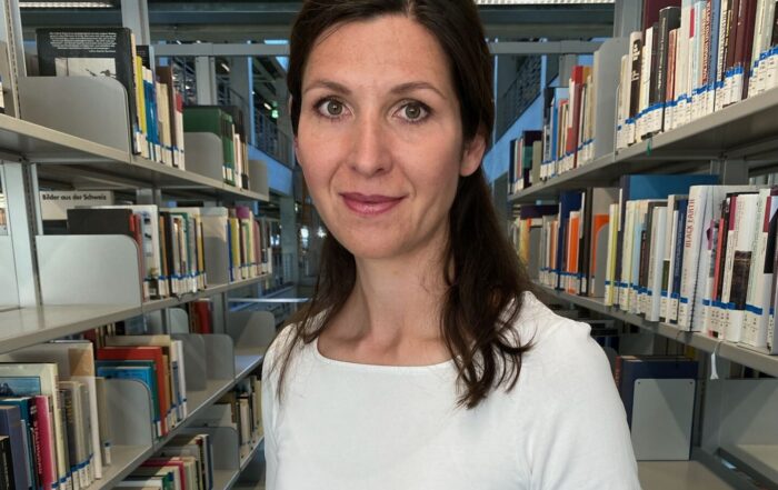 Eine Frau mit langen braunen Haaren steht in einem Gang einer Bibliothek zwischen Regalen voller Bücher. Sie trägt ein weißes Hemd und eine schwarze Uhr und hält ihre Hände zusammen. Die Bibliothek ist modern und großzügig gestaltet. at Hochschule Coburg