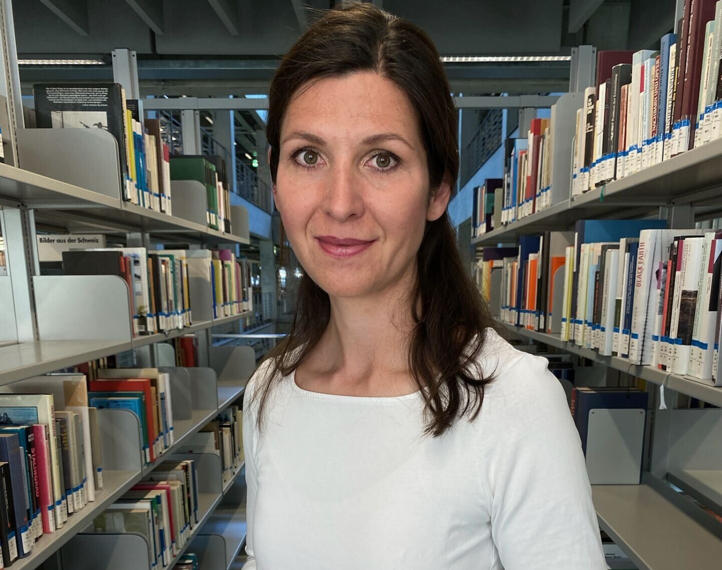 Eine Frau mit langen braunen Haaren steht in einem Gang einer Bibliothek zwischen Regalen voller Bücher. Sie trägt ein weißes Hemd und eine schwarze Uhr und hält ihre Hände zusammen. Die Bibliothek ist modern und großzügig gestaltet. at Hochschule Coburg