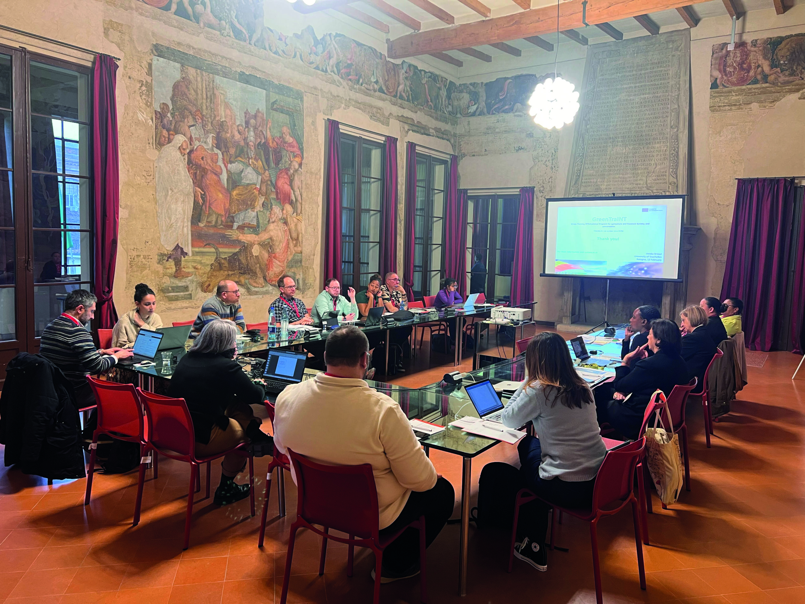 In einem Konferenzraum mit kunstvollen Gemälden und gefliesten Böden sitzt eine Gruppe von Menschen mit Laptops und Papieren in der Hand um Tische. Auf dem Bildschirm an der Vorderseite des Raums läuft eine Präsentation über nachhaltige Landwirtschaft auf den Seychellen. at Hochschule Coburg