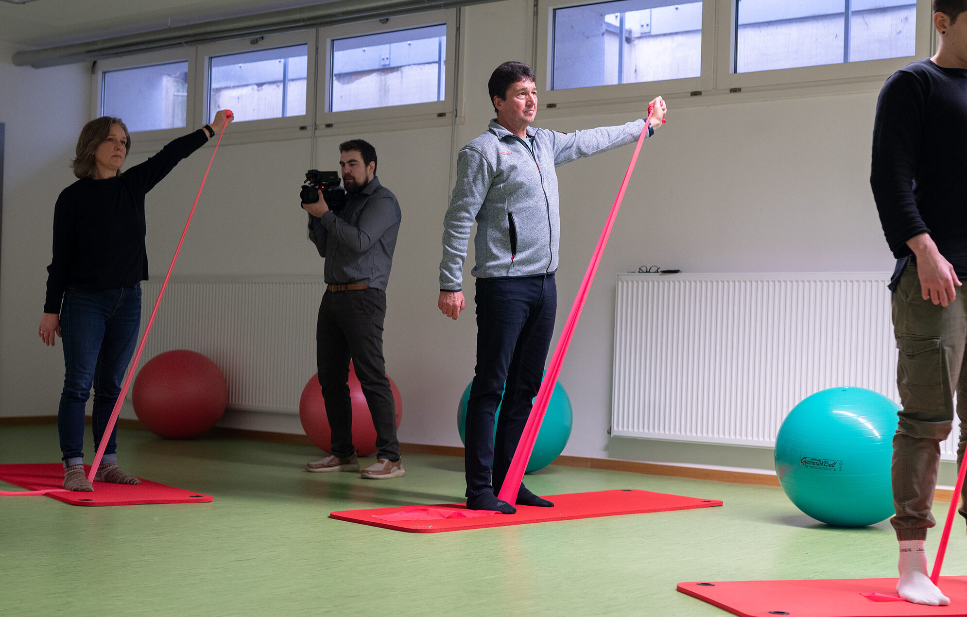 Eine Gruppe von Menschen macht in einem Raum mit großen Fenstern auf roten Matten Gesundheitsförderungsübungen und nutzt dazu rote Widerstandsbänder. Eine Person mit einer Kamera fängt die Szene ein, in der im Hintergrund zwei große Gymnastikbälle liegen. at Hochschule Coburg