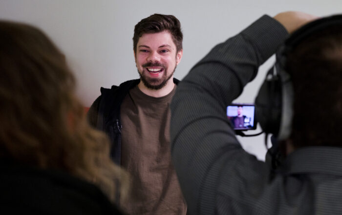 Ein Mann mit Bart lächelt in die Kamera und strahlt Selbstbewusstsein aus. Er trägt ein braunes Hemd und eine schwarze Jacke. at Hochschule Coburg