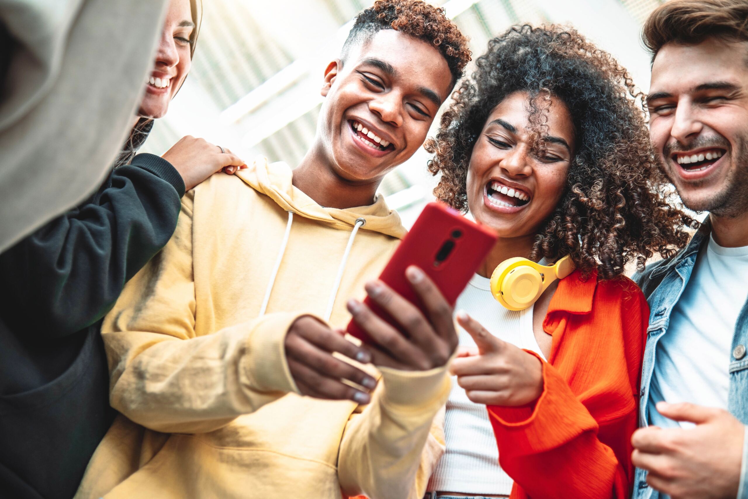 Eine Gruppe von vier Freunden lacht und lächelt und schaut dabei auf ein Smartphone. Sie sind leger gekleidet, eine Person trägt gelbe Kopfhörer um den Hals. Die Stimmung ist fröhlich und entspannt. at Hochschule Coburg