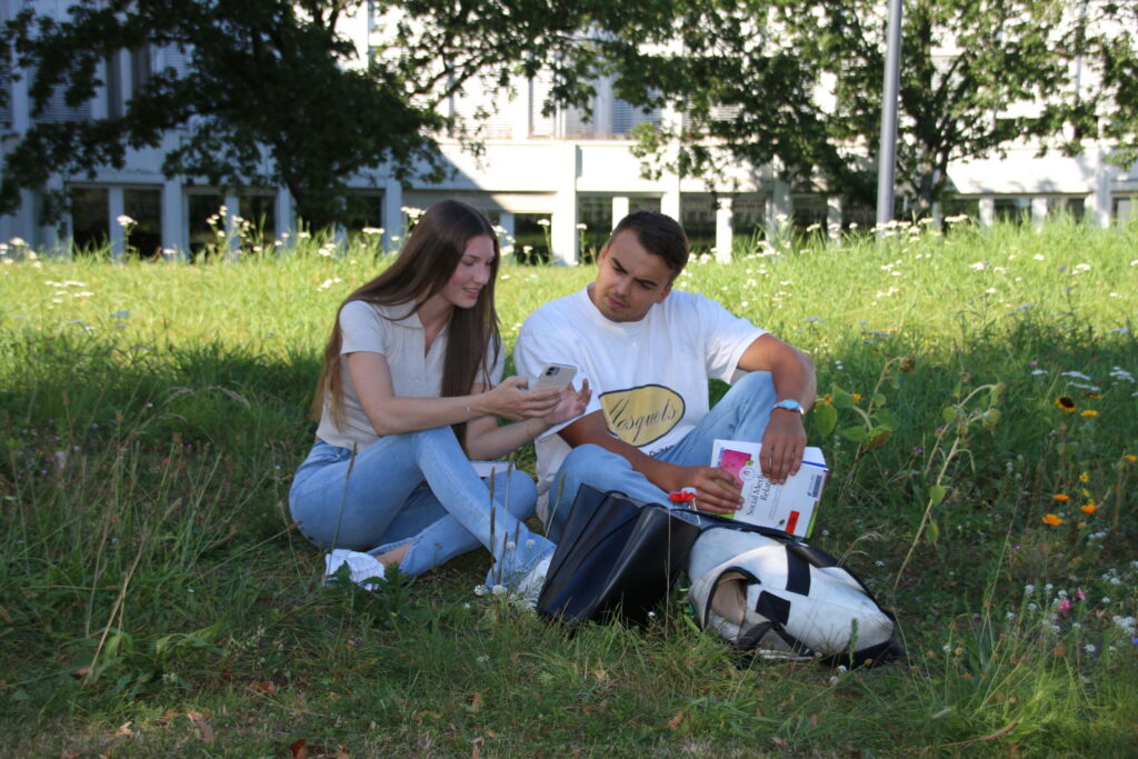 Two people are sitting on the grass, immersed in studying as they share a phone screen. One wears a white shirt and holds a laptop, while the other sports a gray shirt and blue jeans. Backpacks and books are scattered nearby, with a building and trees providing a serene backdrop. at Hochschule Coburg