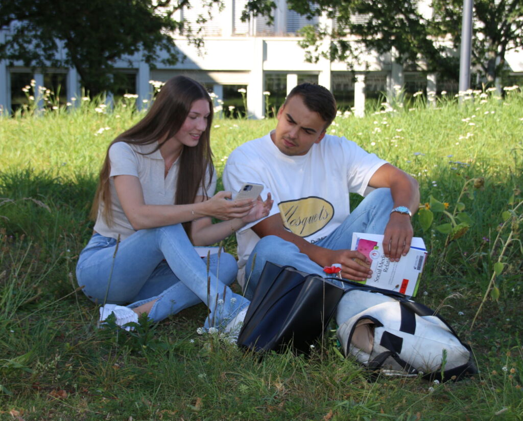 Zwei Personen sitzen auf dem Rasen und sind in ihr Telefon vertieft, während eine von ihnen Bücher zu ihrem Studium auf dem Schoß balanciert. Umgeben von üppigem Grün und bunten Blumen mit einem Gebäude im Hintergrund sind sie leger gekleidet und scheinen sich auf das gemeinsame Lernen zu konzentrieren. at Hochschule Coburg