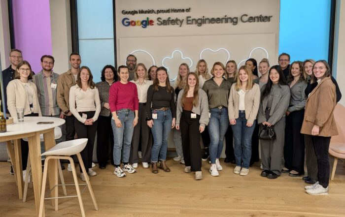 Eine Gruppe von Menschen steht lächelnd vor dem Schild „Google Safety Engineering Center“. Sie sind drinnen, umgeben von Stühlen und einem hohen Tisch, was Googles Vision für die Zukunft der Arbeit in einem Büroumfeld eines Technologieunternehmens widerspiegelt. at Hochschule Coburg