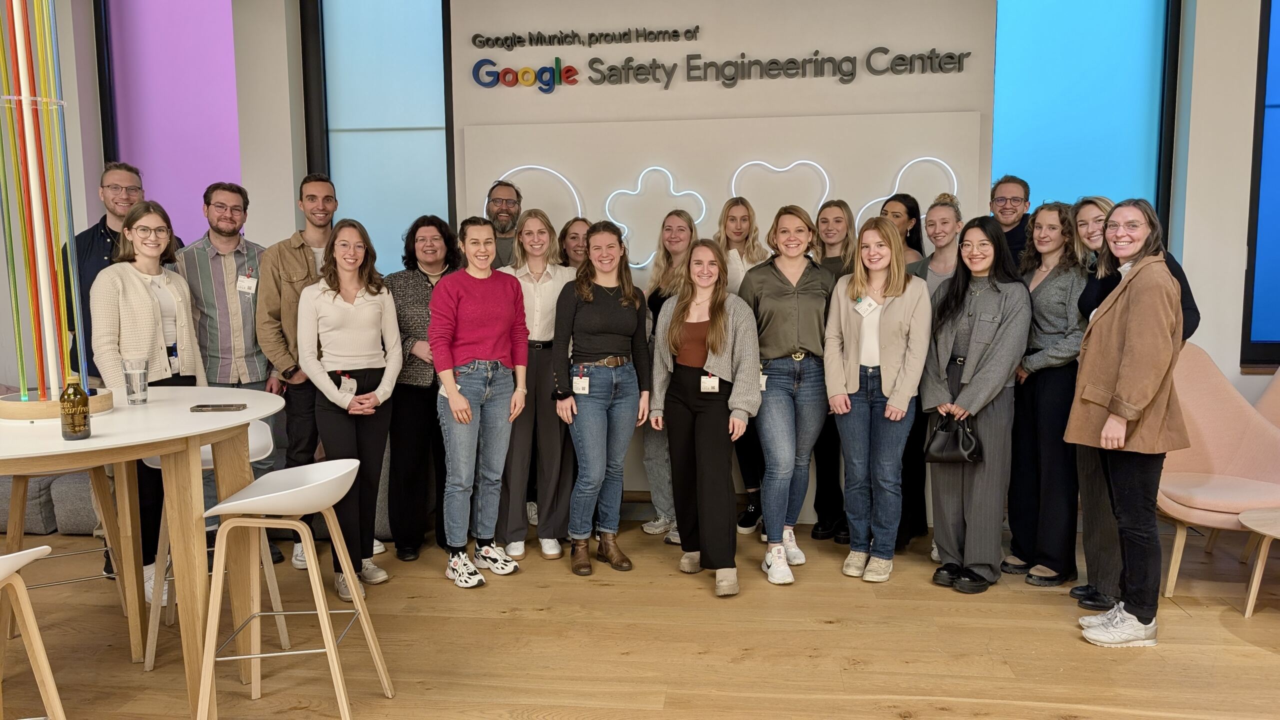 Eine Gruppe von Menschen steht lächelnd vor dem Schild „Google Safety Engineering Center“. Sie sind drinnen, umgeben von Stühlen und einem hohen Tisch, was Googles Vision für die Zukunft der Arbeit in einem Büroumfeld eines Technologieunternehmens widerspiegelt. at Hochschule Coburg