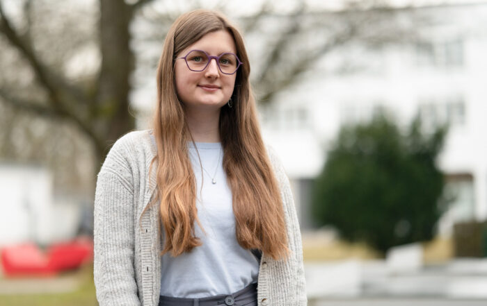 Eine junge Person mit langen braunen Haaren und Brille steht im Freien und trägt einen hellgrauen Pullover über einem blauen Hemd. Die Szene hat eine entwurfsähnliche Qualität, da der unscharfe Hintergrund kunstvoll einen Baum und Gebäude darstellt. at Hochschule Coburg