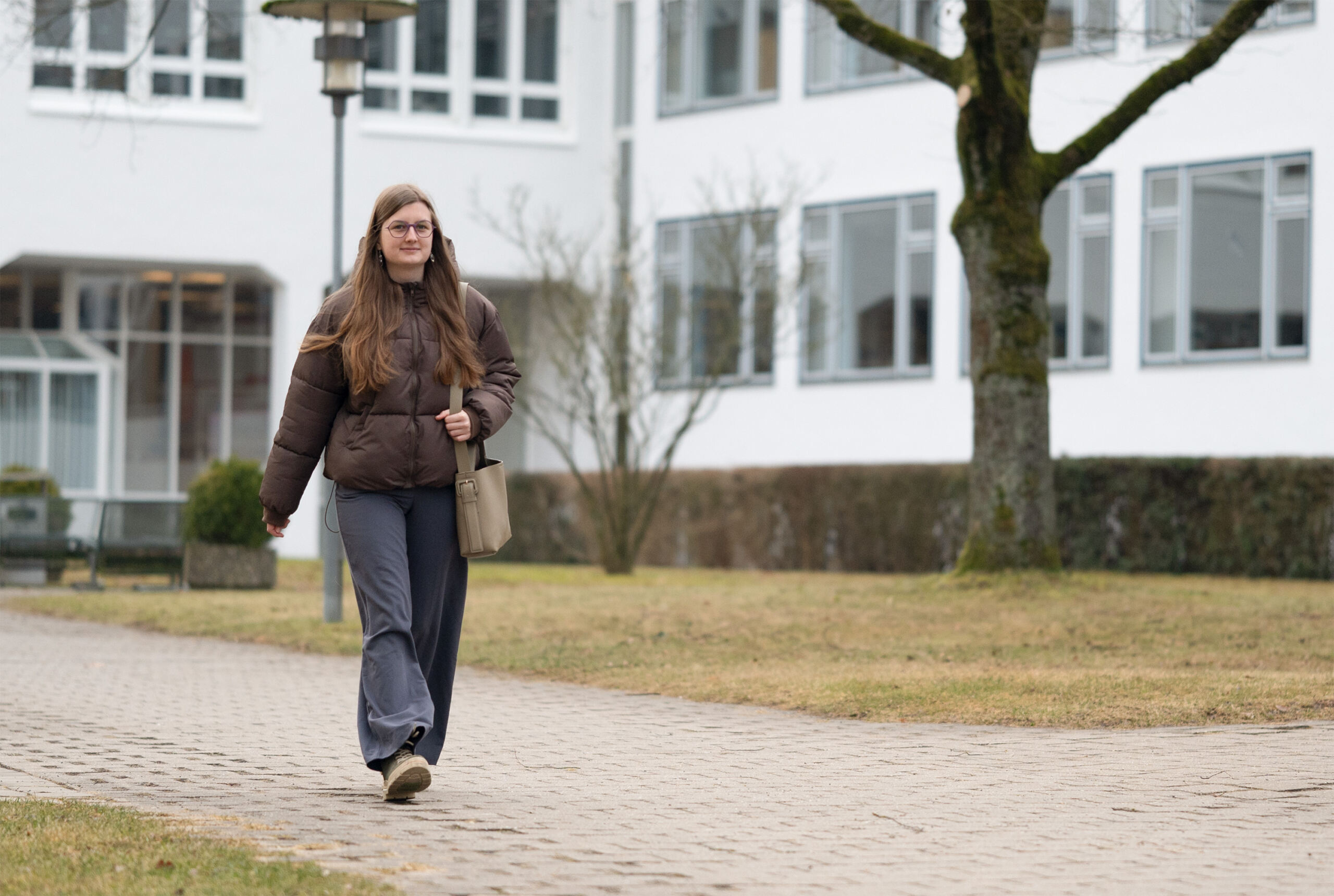 Eine Person mit langen Haaren und Brille schlendert einen gepflasterten Weg in einem Wohngebiet entlang und gleicht einem „automatisch gespeicherten Entwurf“ städtischer Ruhe. Sie trägt eine braune Jacke, graue Hosen und eine Tasche. Im Hintergrund sind Bäume und weiße Gebäude mit großen Fenstern zu sehen. at Hochschule Coburg