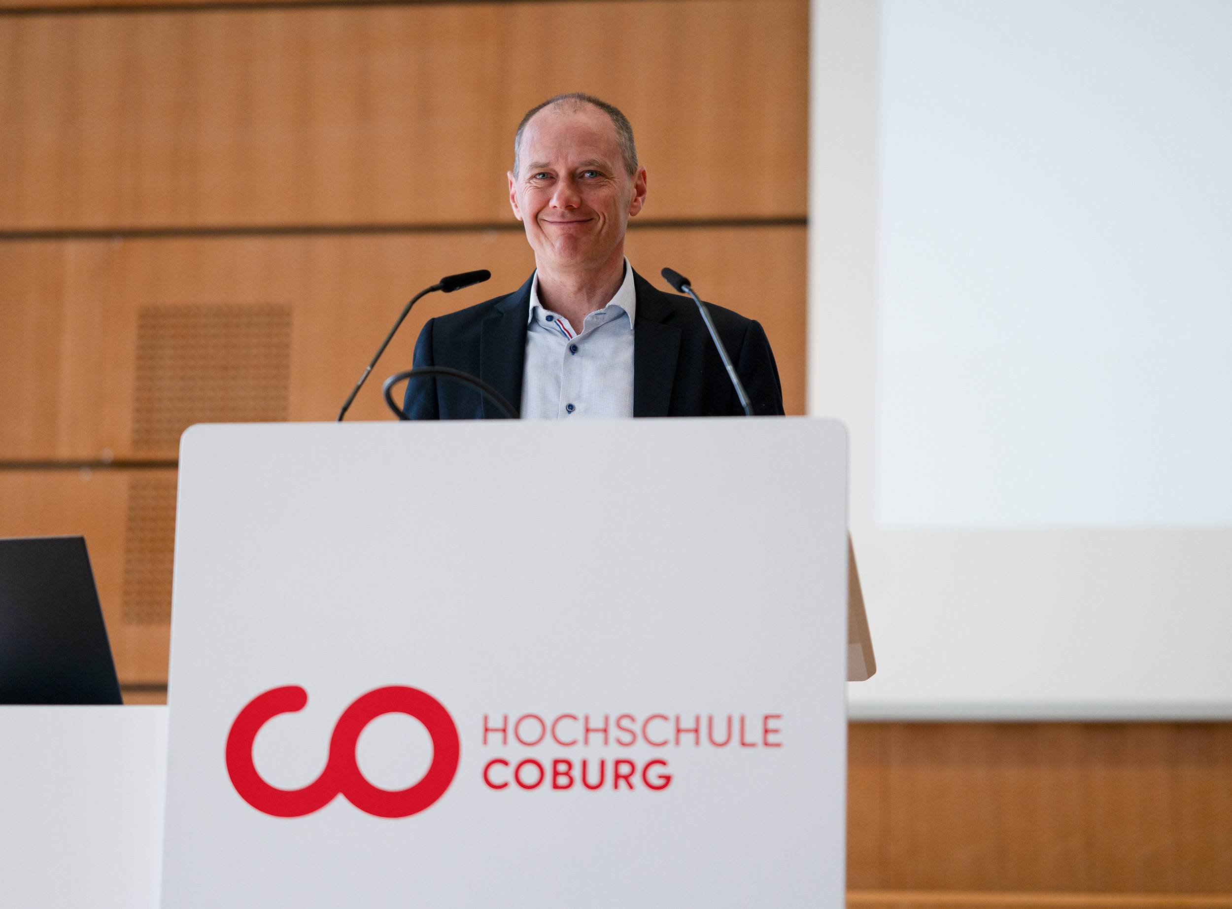 Ein Mann im Anzug, Fachrichtung Soziale Arbeit, steht lächelnd an einem Podium mit der Aufschrift „Hochschule Coburg“ in einem holzgetäfelten Raum. Hinter ihm ist ein weißer Bildschirm, zwei Mikrofone ragen auf ihn zu. at Hochschule Coburg