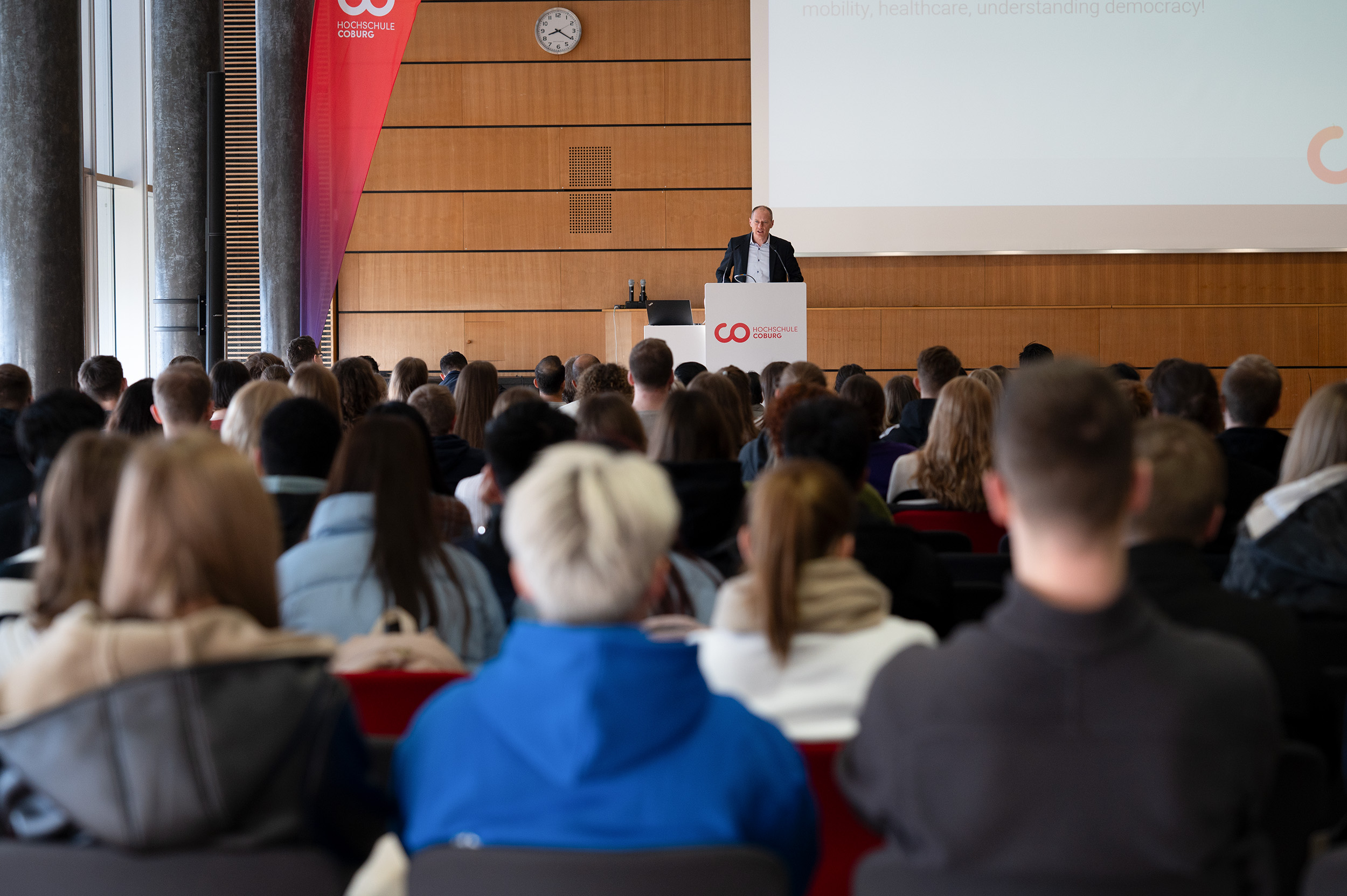 Ein Redner steht an einem Podium und spricht vor einem großen Publikum in einem Auditorium über Internationales Finanzmanagement. Die Anwesenden verfolgen aufmerksam die Präsentation. Hinter dem Redner sind ein rot-weißes Banner und eine Wanduhr zu sehen. at Hochschule Coburg