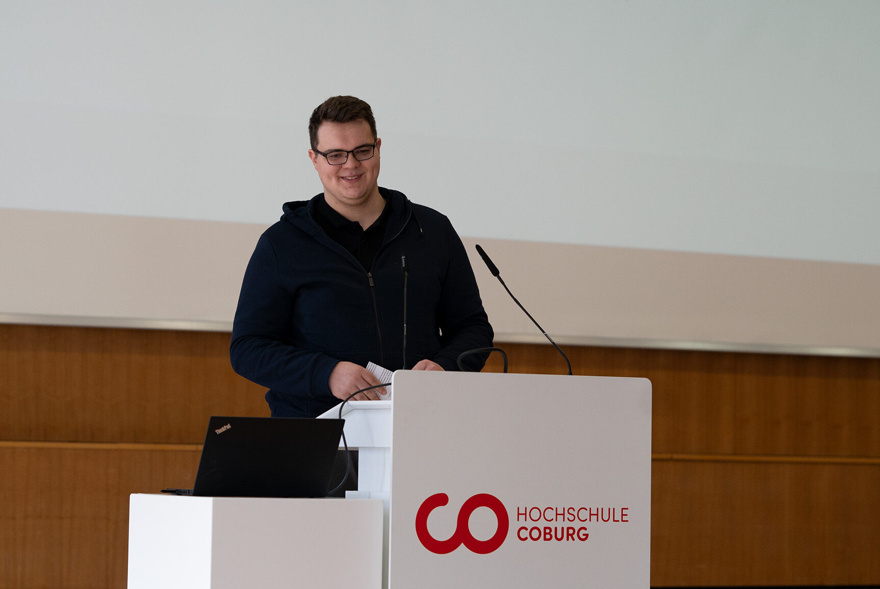 Eine Person in dunkler Jacke steht an einem Podium und hält einen Vortrag. Das Podium zeigt das Logo der Hochschule Coburg. Im Hintergrund wird eine Folie projiziert, auf der „Jan Keller“ und „Studentenvertretung“ zu sehen sind. Sie diskutieren Aspekte des Internationalen Finanzmanagements. at Hochschule Coburg