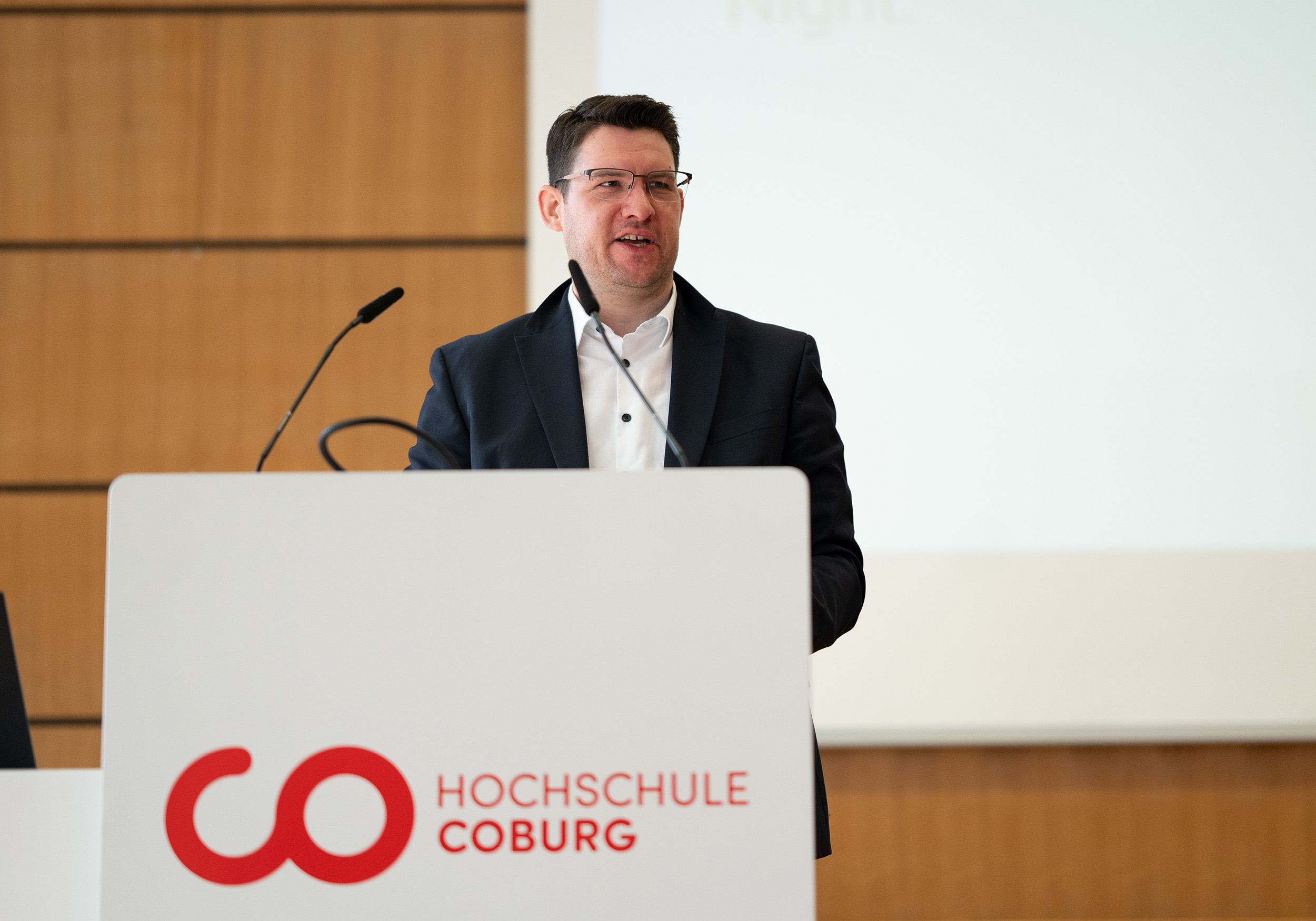 Ein Mann in dunklem Anzug und Brille steht an einem weißen Podium mit Mikrofon und spricht. Anlässlich des Semesterstarts der Hochschule Coburg trägt das Podium das Logo der Hochschule. Im Hintergrund sind eine Holzpaneelwand und eine Projektionsleinwand zu sehen, die die Bühne für Diskussionen zum Thema Soziale Arbeit bilden. at Hochschule Coburg