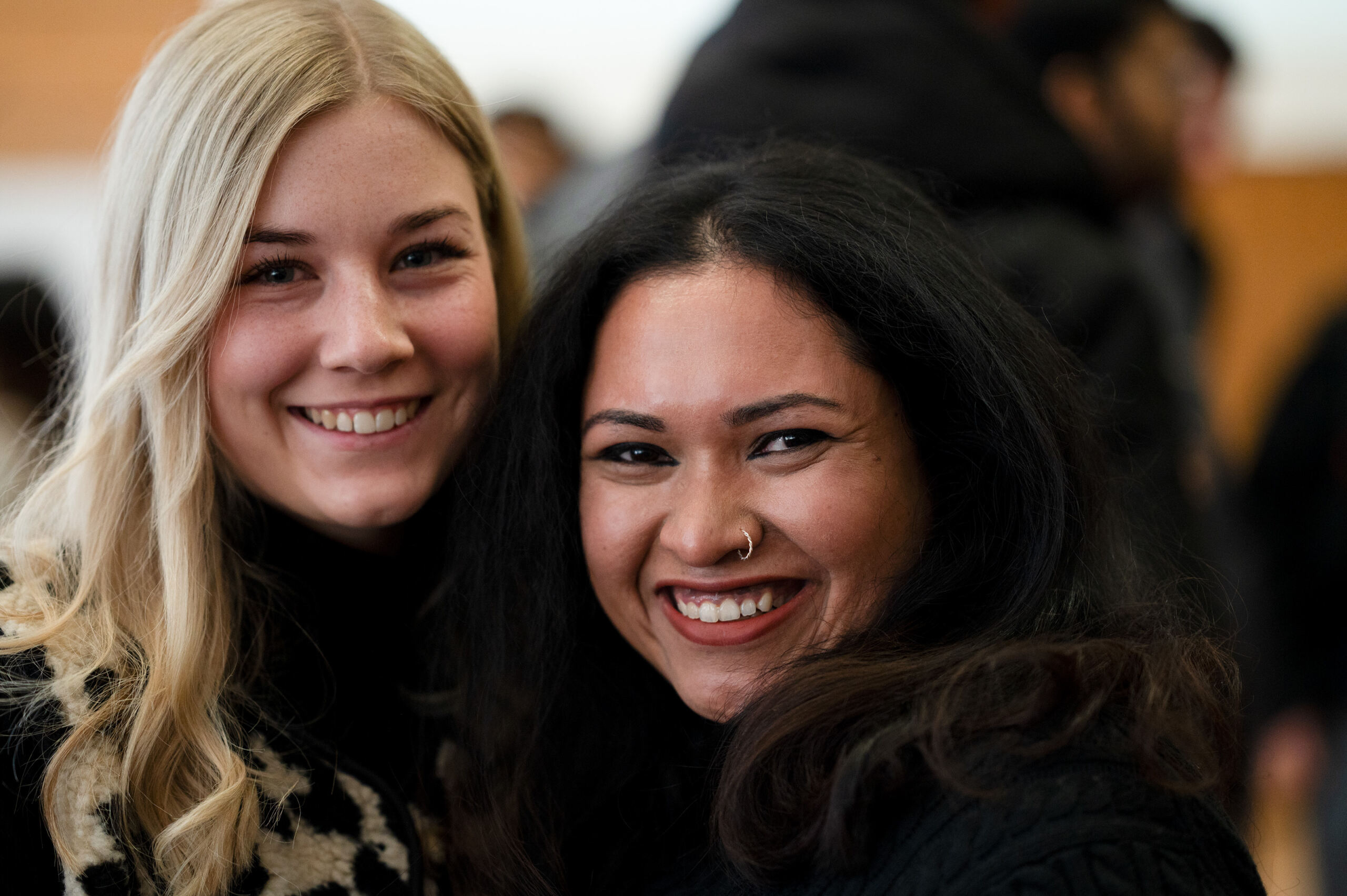 Zwei Personen stehen lächelnd und dicht beieinander in einem Raum. Die linke Person hat lange blonde Haare, die rechte lange dunkle Haare und einen Nasenring. Beide tragen dunkle Kleidung vor einem leicht verschwommenen Hintergrund mit anderen Personen. at Hochschule Coburg