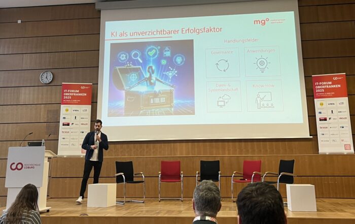Ein Redner steht auf einer Bühne in einem Konferenzraum und präsentiert Folien zum Thema KI. Im Publikum sitzen einige Personen. Die Folie enthält Grafiken und Text. Der Raum ist modern gestaltet und verfügt über Holzvertäfelungen und ein großes Fenster. at Hochschule Coburg