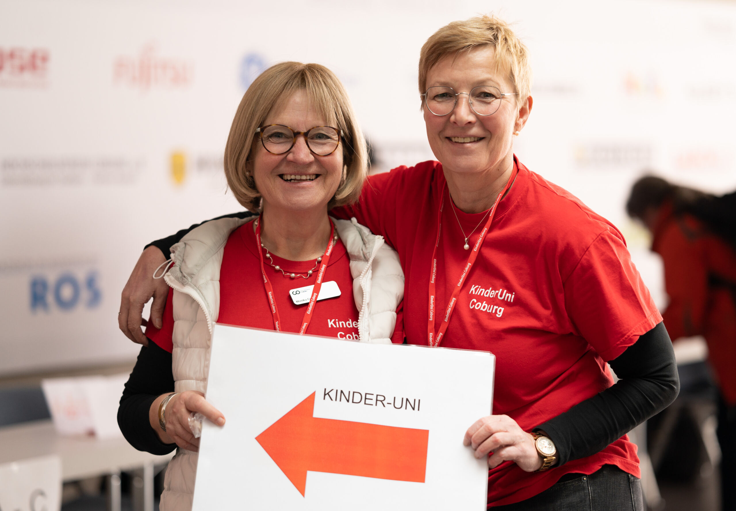 Zwei lächelnde Personen in roten Shirts mit der Aufschrift „Kinder-Uni“ halten ein Schild mit einem großen Pfeil und dem Text „KINDER-UNI“. Sie stehen vor einem unscharfen Hintergrund mit verschiedenen Logos. at Hochschule Coburg