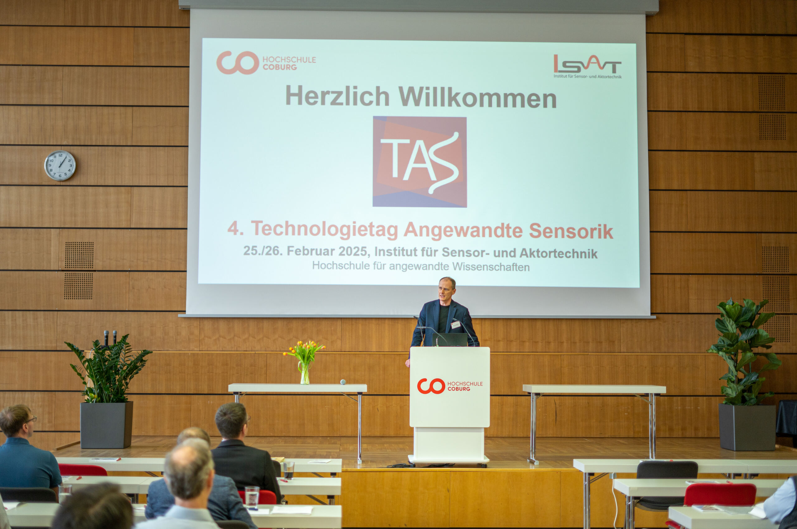 Ein Mann steht an einem Rednerpult in einem Konferenzraum und spricht zu seinen Zuhörern. Hinter ihm begrüßt ein Bildschirm die Teilnehmer zum „4. Technologietag Angewandte Sensorik“ und stellt Fortschritte in der Medizintechnik vor. Der Raum ist mit Holzvertäfelungen und Topfpflanzen ausgestattet, was eine einladende Atmosphäre schafft. at Hochschule Coburg