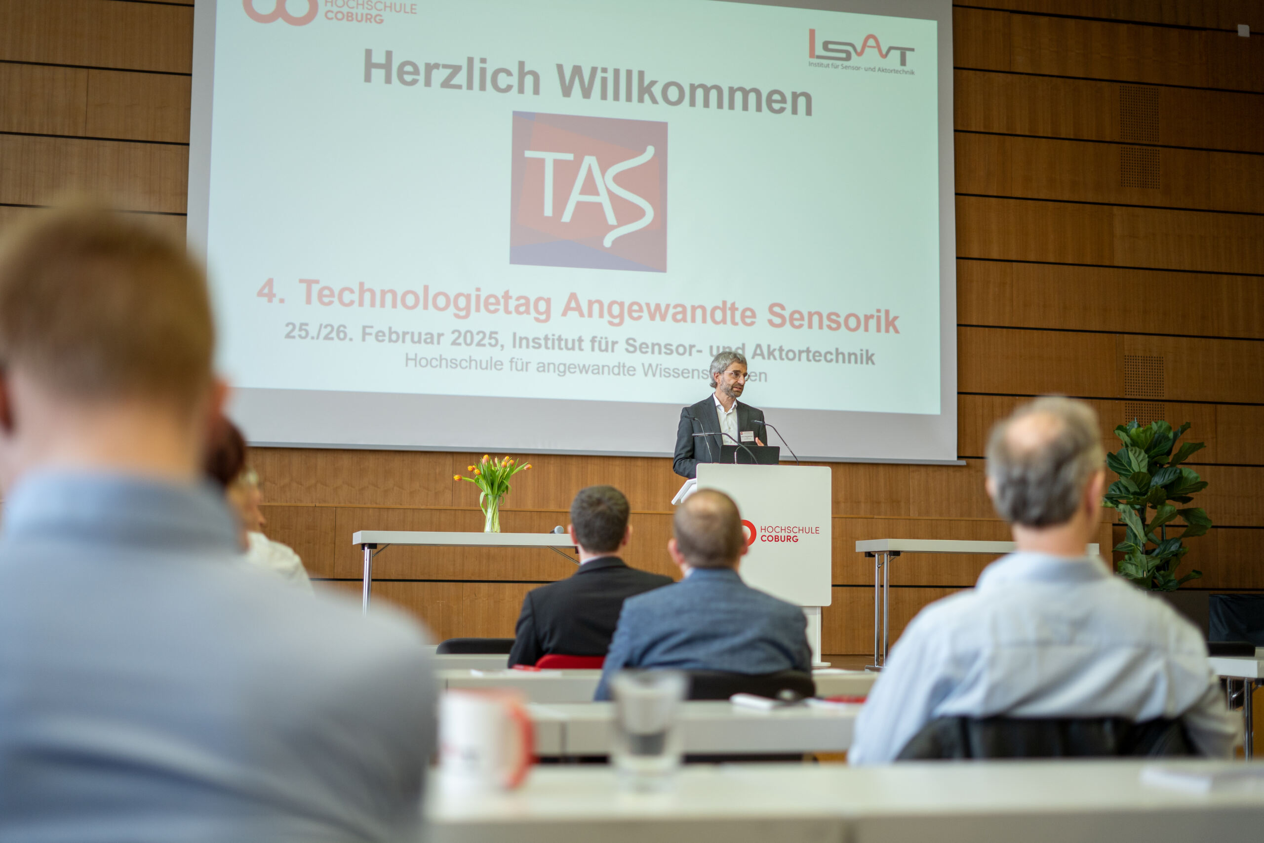 Ein Redner steht an einem Podium und spricht zu einem Publikum in einem Konferenzraum. Auf einem großen Bildschirm dahinter wird eine Begrüßungsansprache für den 4. Technologietag Angewandte Sensorik angezeigt, bei der Innovationen in der Medizintechnik hervorgehoben werden. Die Teilnehmer sitzen und blicken dem Redner aufmerksam zu. at Hochschule Coburg