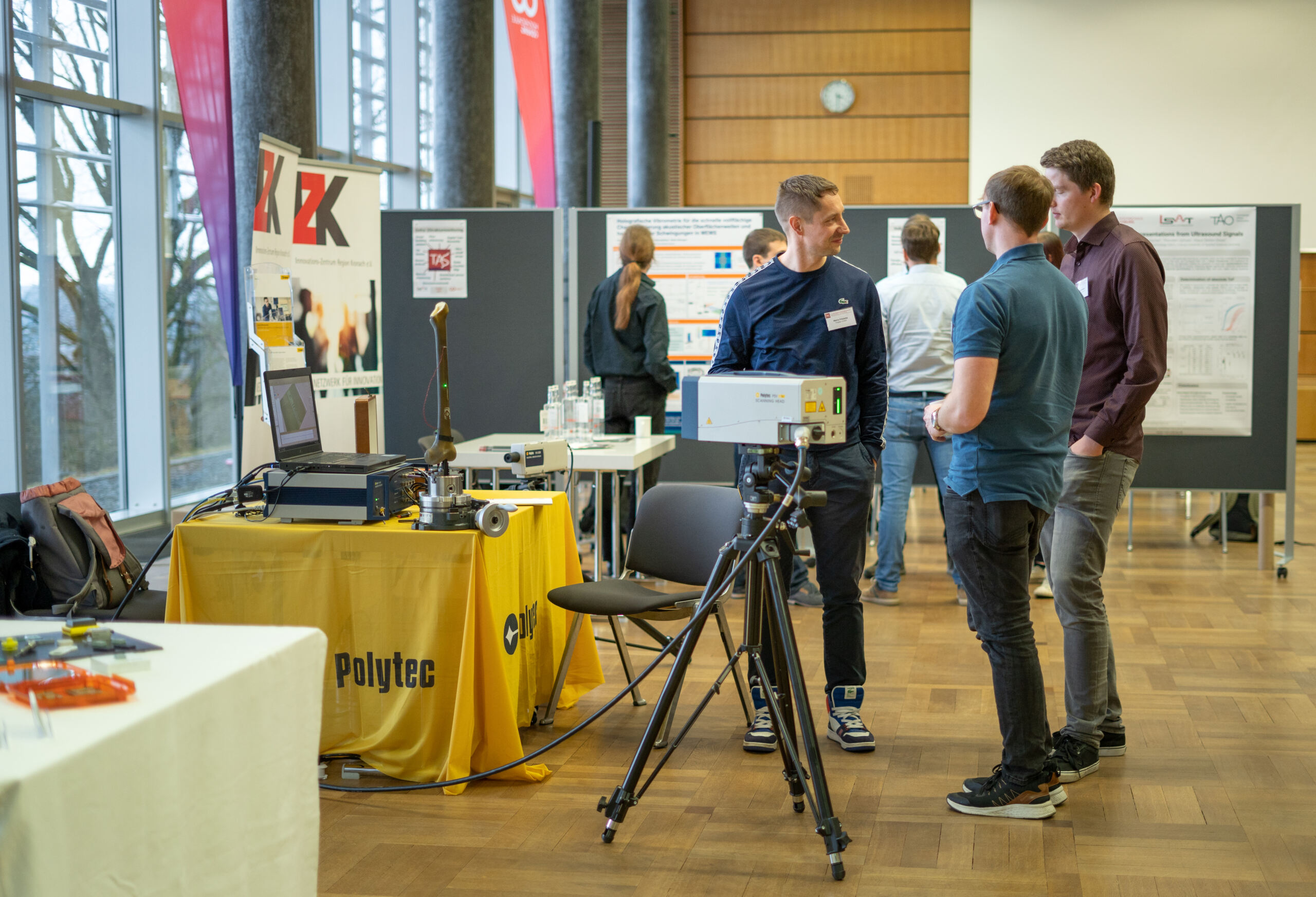 Drei Personen unterhalten sich neben einem Tisch mit Geräten und einem Polytec-Banner auf einer Technologieausstellung zum Thema Medizintechnik. Im Hintergrund sind Poster und Schautafeln zu sehen, außerdem erkunden andere Besucher den Veranstaltungsort. at Hochschule Coburg