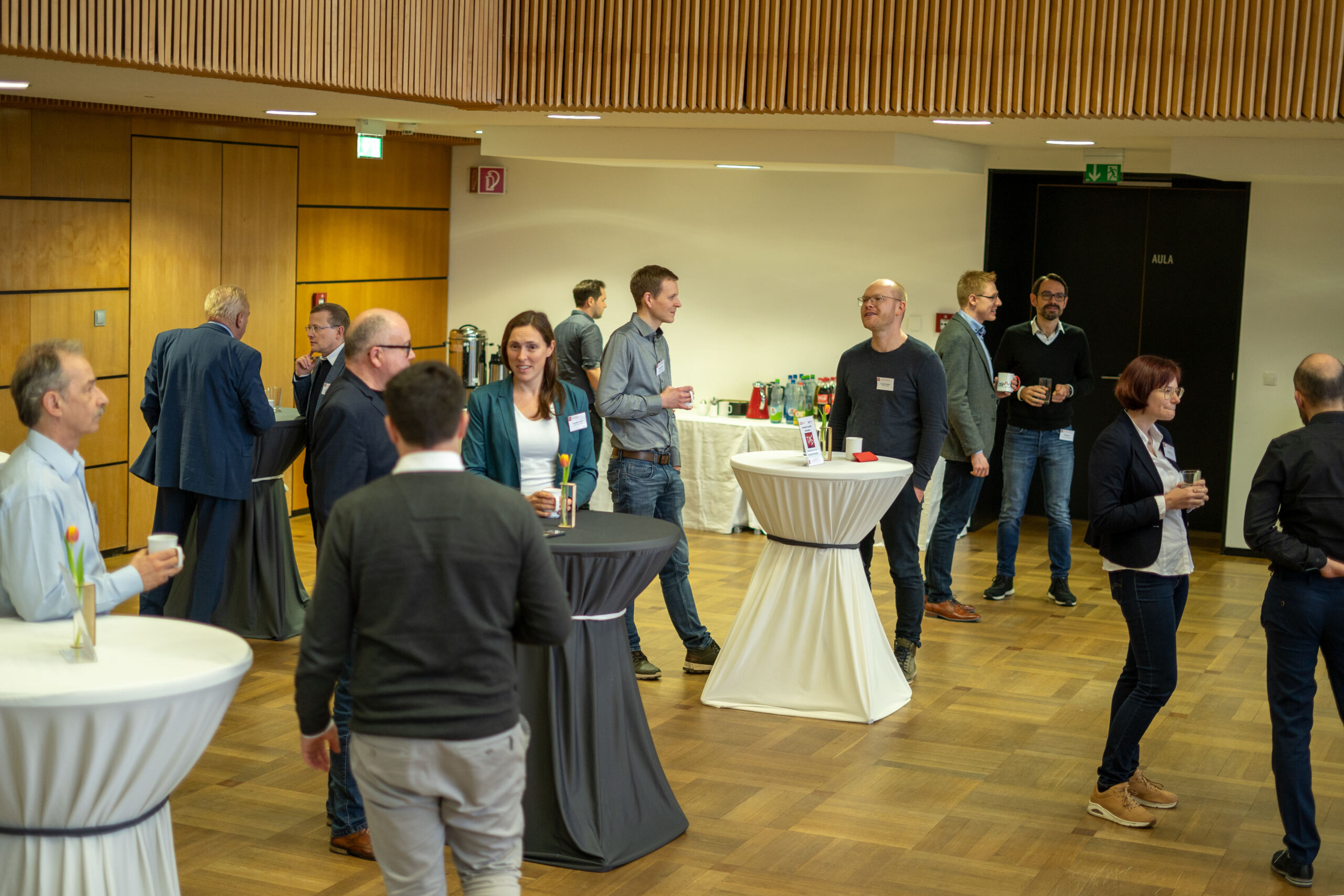 Menschen in legerer Business-Kleidung treffen sich und unterhalten sich bei einer Networking-Veranstaltung zum Thema Recycling in einem modernen, holzgetäfelten Veranstaltungsort. Einige halten Getränke in der Hand und stehen an weißen Cocktailtischen. Im Hintergrund ist ein Tisch mit Erfrischungen zu sehen. at Hochschule Coburg