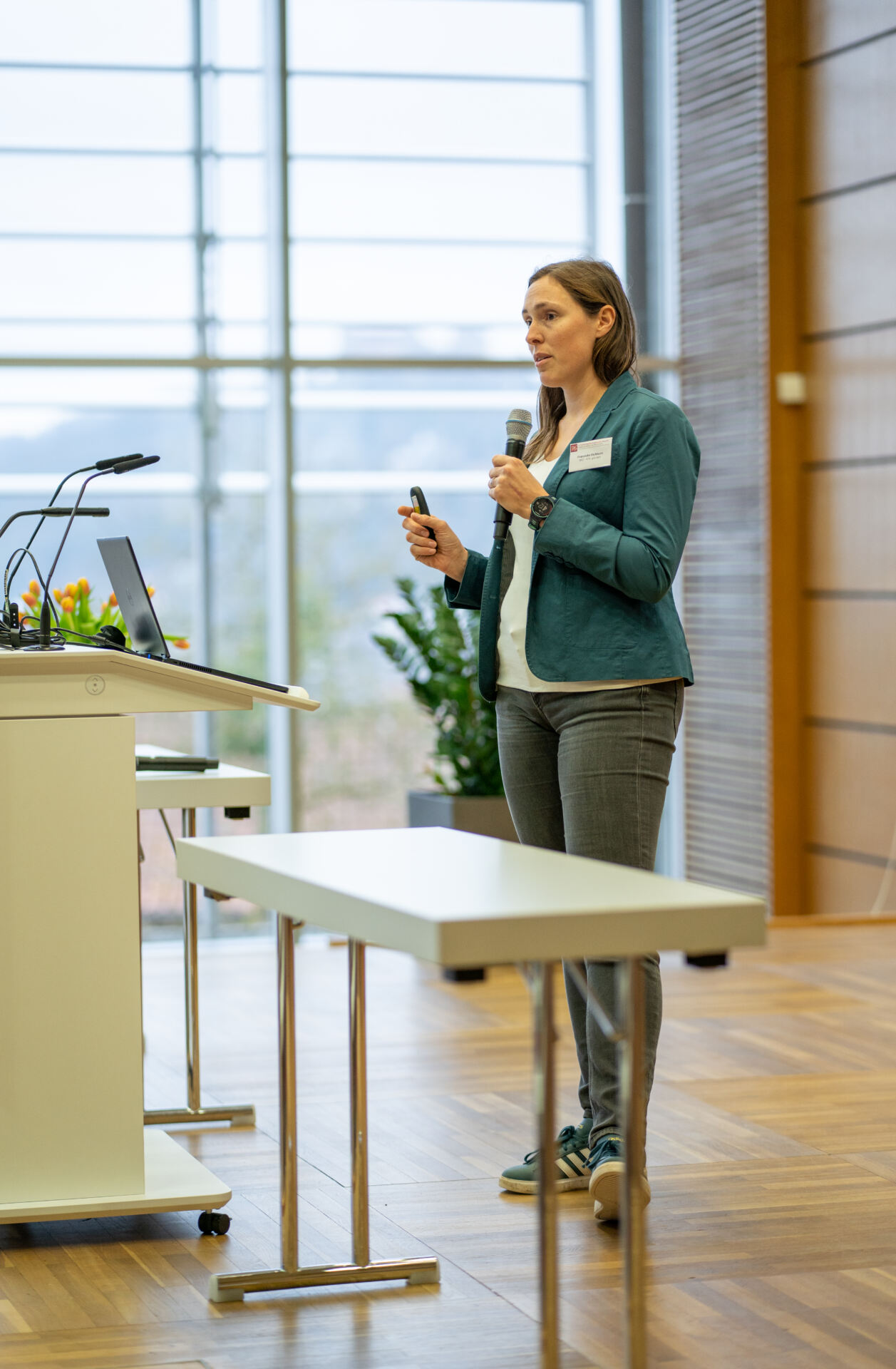 Eine Person in einem grünen Blazer und Jeans spricht in ein Mikrofon, während sie mit einem Laptop neben einem Podium steht und in einem modernen Raum mit großen Fenstern und Pflanzen im Hintergrund eine Präsentation zum Thema Katastrophenschutz hält. at Hochschule Coburg