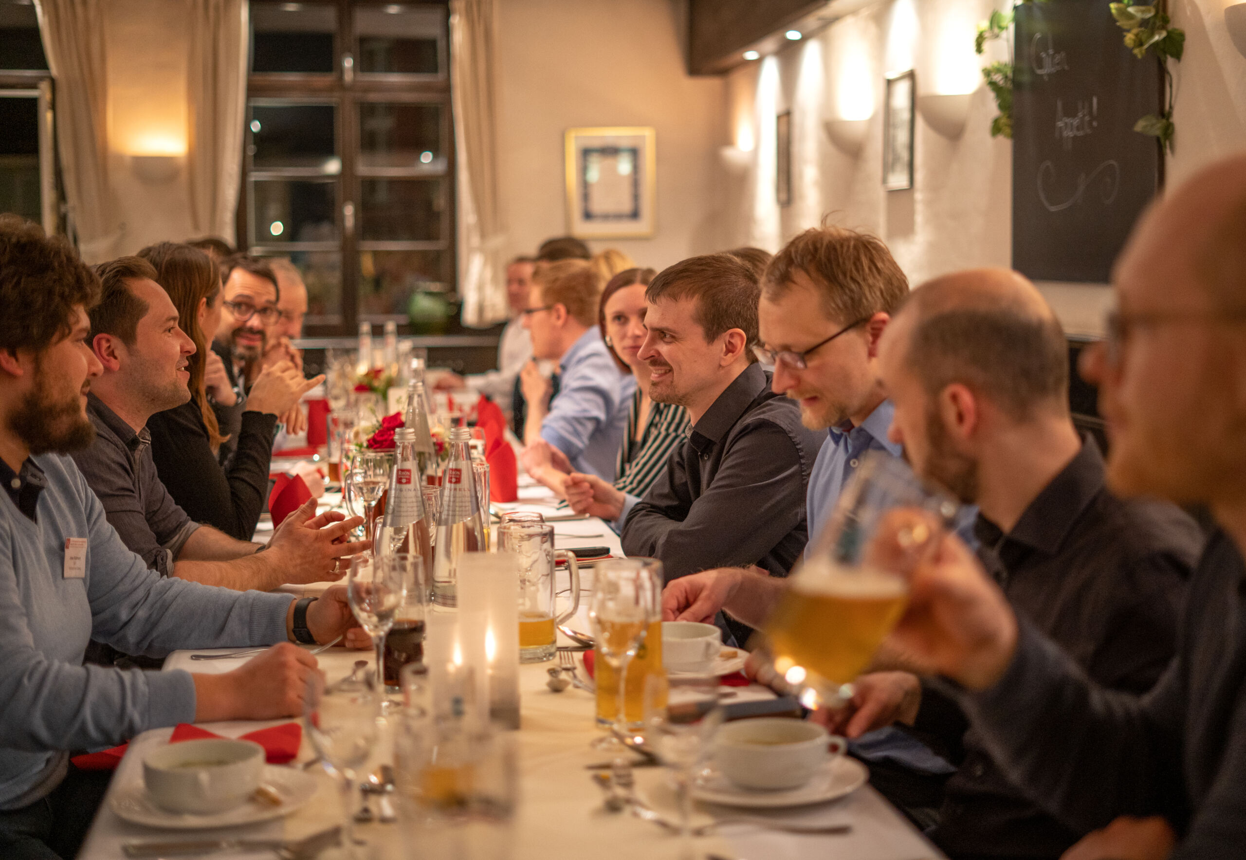 Eine große Gruppe von Menschen sitzt an einem langen Tisch in einem warm beleuchteten Restaurant, unterhält sich und genießt Getränke. Der Tisch, elegant gedeckt mit Gläsern, Flaschen und Kerzen, strahlt eine gemütliche Atmosphäre aus – ein einladender Rückzugsort, der sich perfekt eignet, um über alles zu sprechen, von Sensorik-Innovationen bis hin zu alltäglichen Freuden. at Hochschule Coburg