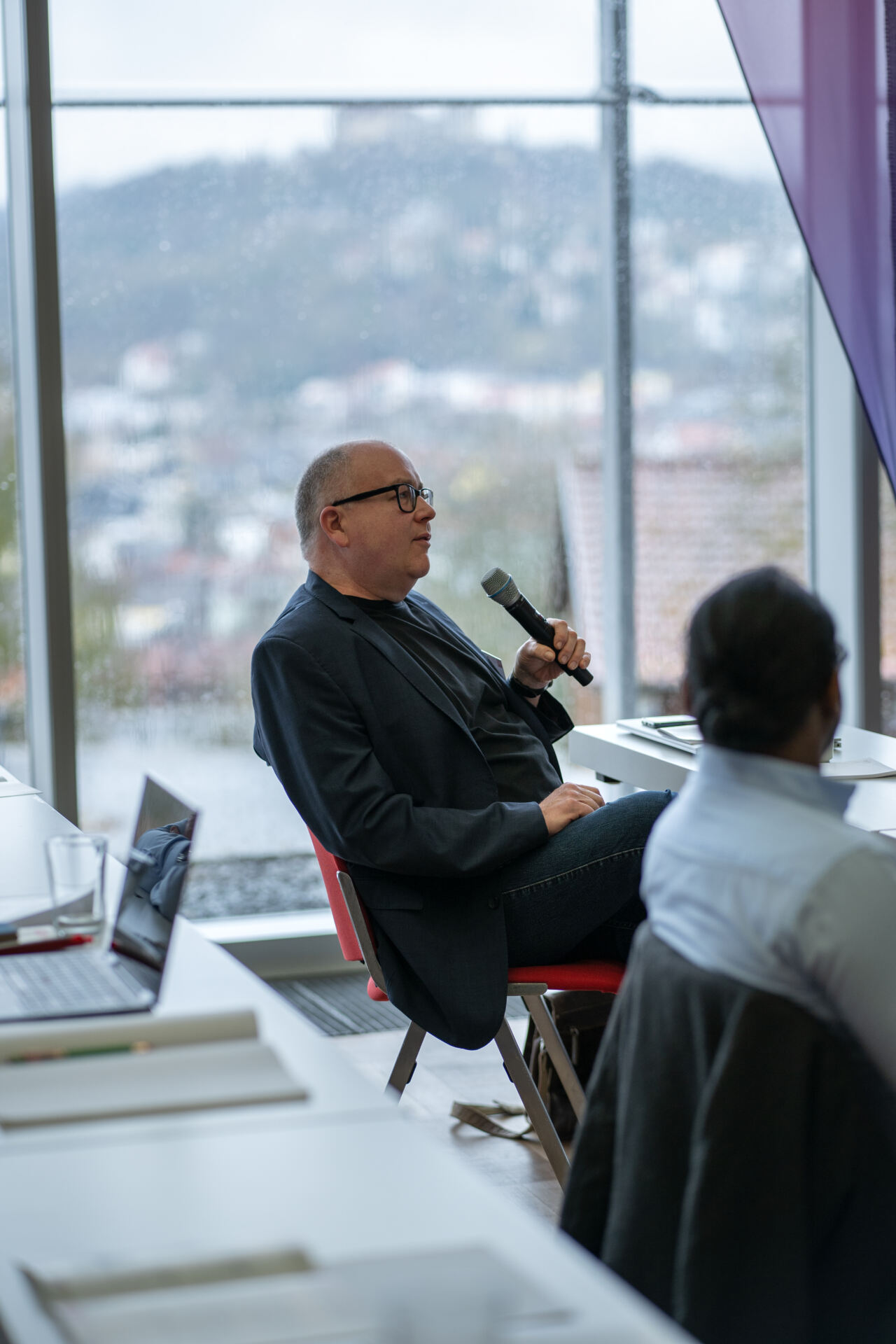 Ein Mann sitzt an einem Tisch mit Laptops und spricht in ein Mikrofon. Er ist in eine Diskussion über Katastrophenschutz vertieft. Er trägt eine Brille und einen dunklen Blazer. In seiner Nähe sitzt eine weitere Person. Durch das große Fenster sind die Stadt und die Berge im Hintergrund zu sehen. at Hochschule Coburg