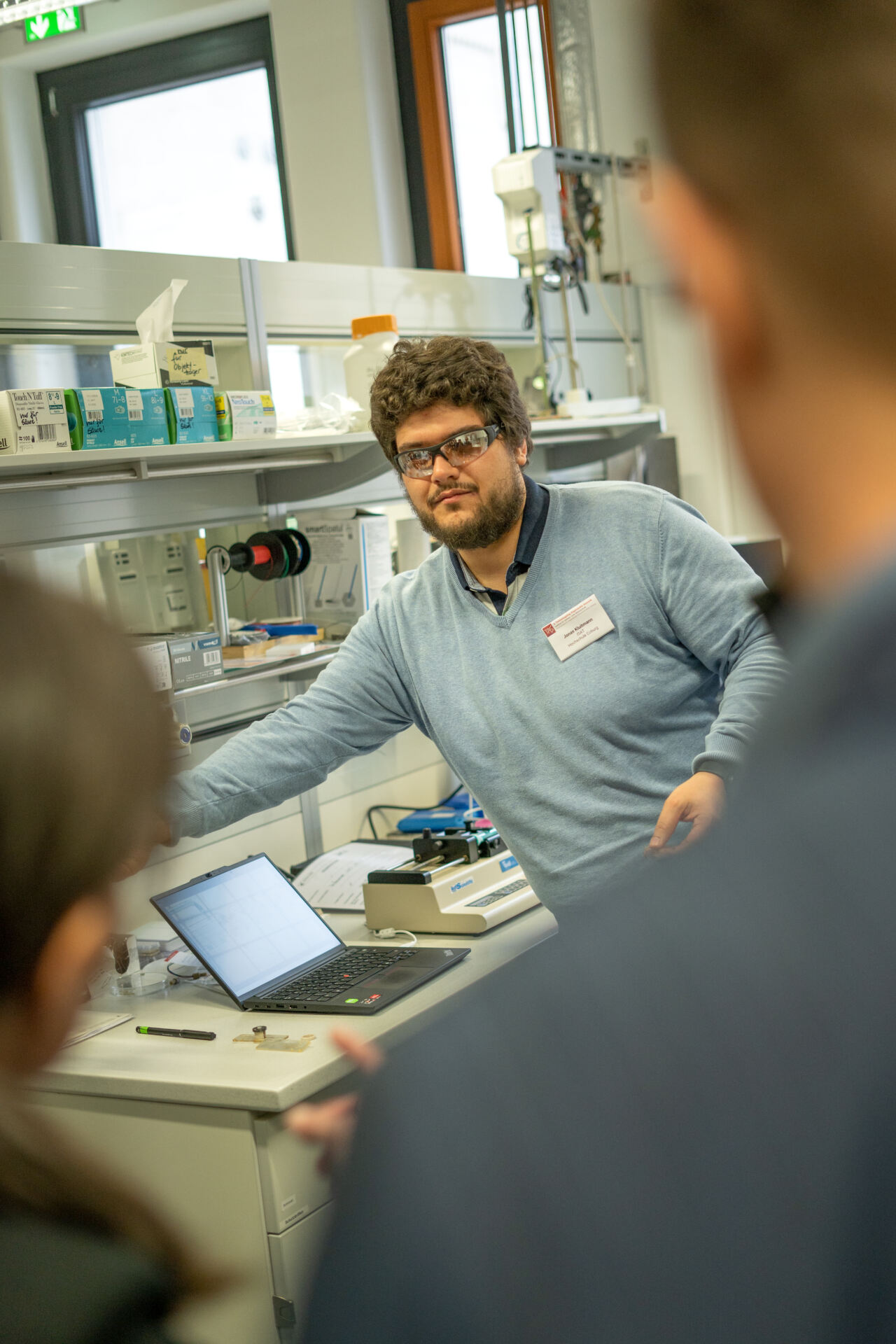 Eine Person mit Brille und hellblauem Pullover steht in einer Laborumgebung und präsentiert innovative Medizintechnik auf einem Tisch mit aufgeklapptem Laptop. Verschwommene Figuren im Vordergrund hören aufmerksam zu. Im Hintergrund sind Regale mit verschiedenen Geräten zu sehen. at Hochschule Coburg